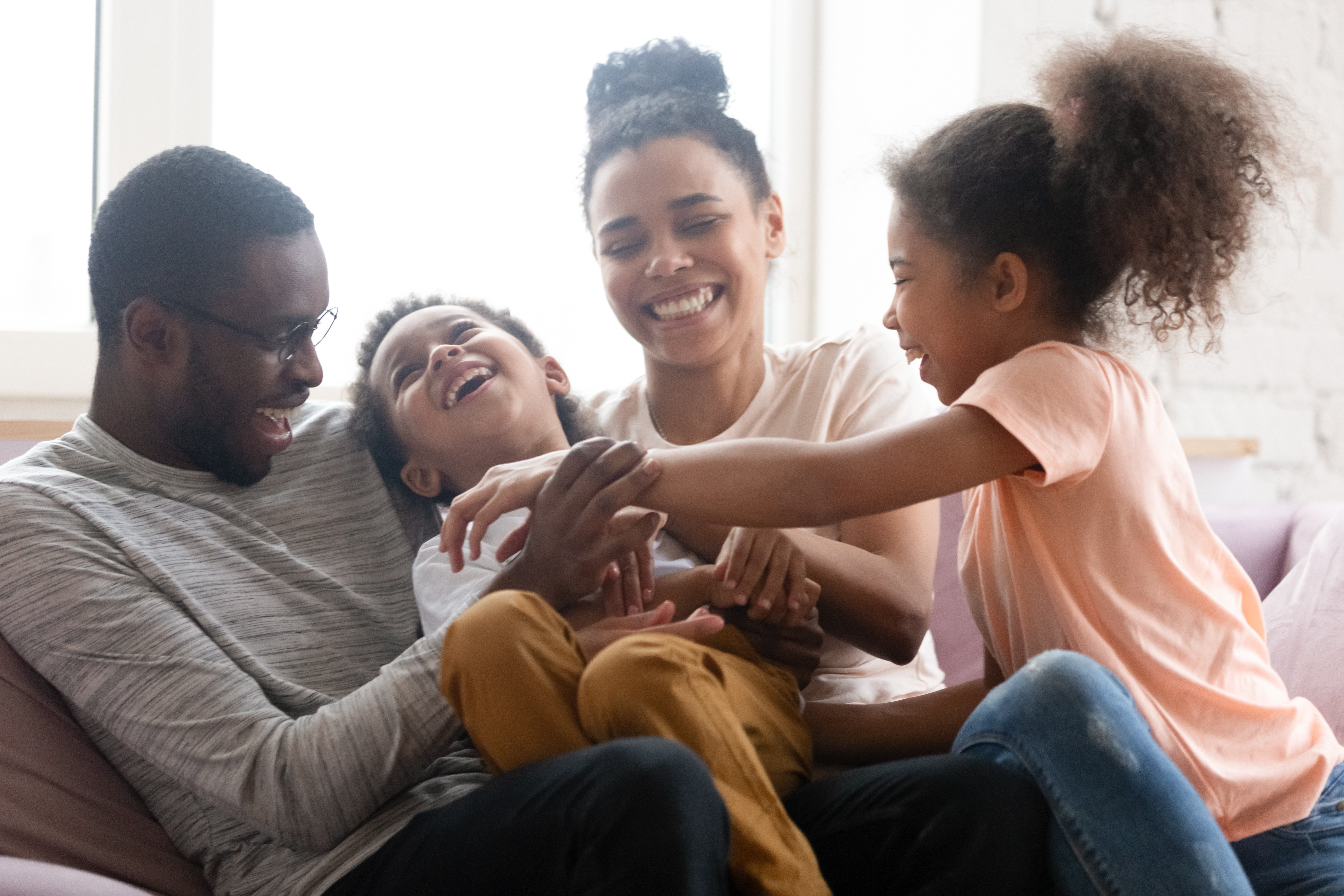 A joyful family enjoying a playful moment together at home, with two parents and two children laughing and interacting on a couch.
