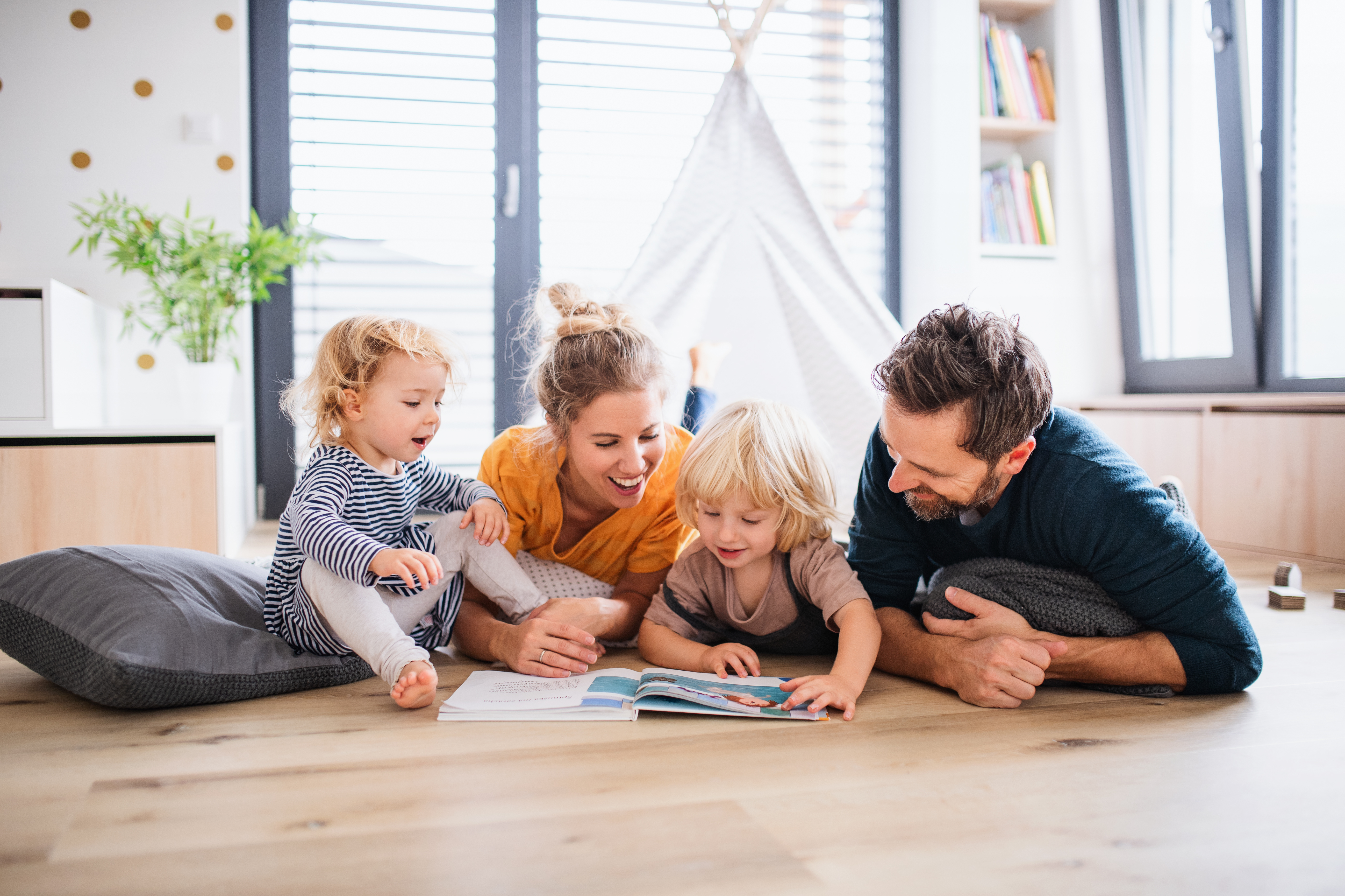 A family of four, including two children, enjoys reading a book together on the floor in a cozy living room. The mother, father, and kids are smiling and engaged, surrounded by cushions and natural light from large windows.