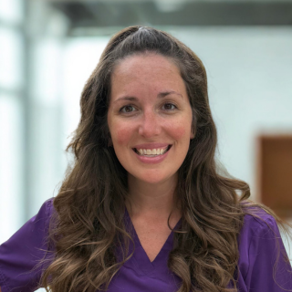 Smiling woman with long, wavy hair wearing a purple scrub top, in a well-lit indoor setting.